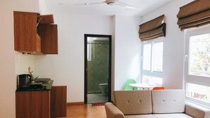 Un ambiente compatto con un divano, un mobiletto in legno, una ventola a soffitto, una finestra con tende e una porta che conduce al bagno.
