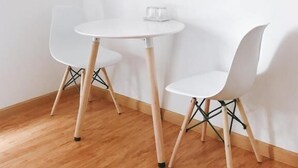 Un piccolo tavolo rotondo bianco con due sedie bianche. Sul tavolo c'è un bicchiere. Le sedie hanno gambe in legno e struttura in metallo nero. Il pavimento è in legno.