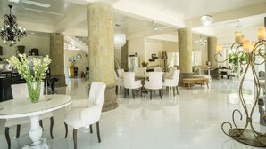 Un amplio y luminoso vestíbulo de hotel con zona de comedor central, grandes columnas y modernas lámparas de araña.