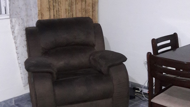 A living room with a brown recliner, a wooden dining table with chairs, a patterned curtain, and a tiled floor. - Oaklands Apartment complex (Kingston)