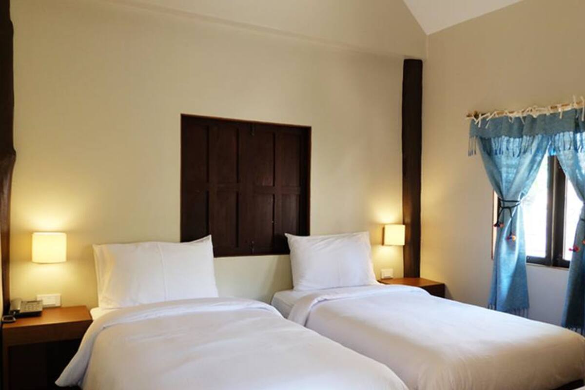A hotel room with two single beds, wooden headboard, bedside lamps, and a window with blue curtains.