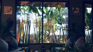 A room with a large window overlooking a garden with tall plants and a wooden fence. There are two wall-mounted artworks and a table with a vase of flowers. - DaNang Moon Villa & Apartment (Da Nang)