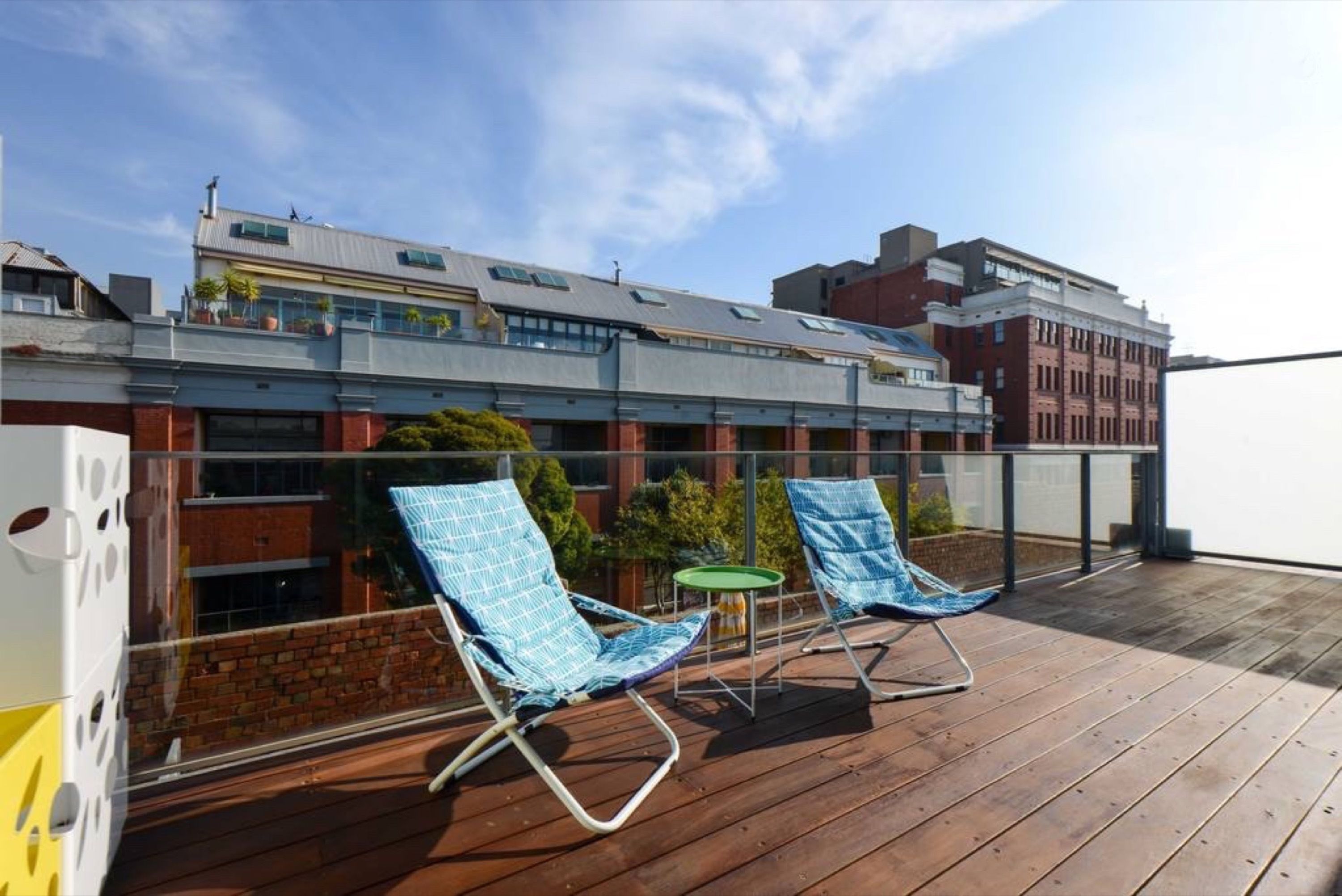 A rooftop terrace with two blue lounge chairs and a small table, overlooking a cityscape with modern buildings.