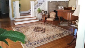 A spacious living room with hardwood floors, a large patterned rug, a piano, and a staircase leading to another level.