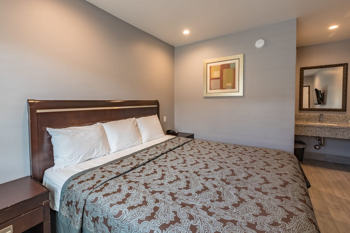 Uma cama bem arrumada com colcha estampada, uma mesinha de cabeceira, um espelho fixado na parede e um banheiro com bancada de granito.