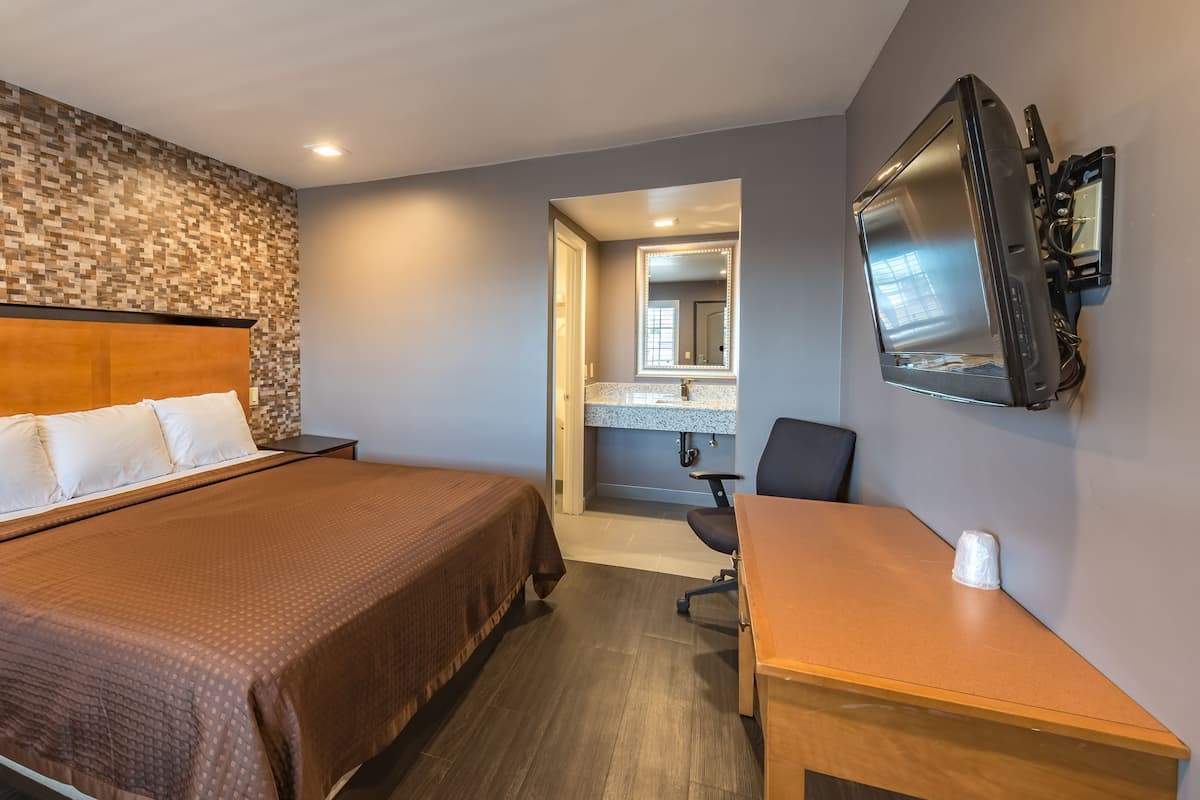 Quarto de hotel com cama, escrivaninha, cadeira, televisão e banheiro com espelho.