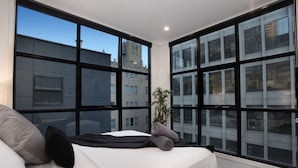Un dormitorio moderno con una cama grande, una planta en maceta y vistas a edificios a través de amplios ventanales.