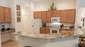 A kitchen with wooden cabinets, a white refrigerator, a microwave, an oven, a granite countertop, and two bar stools. - Sm215701 - Watersong - 5 Bed 4 Baths Villa (Davenport)