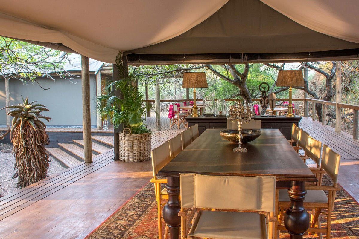 A covered dining area with a long table and chairs, surrounded by potted plants and a wooden bench.