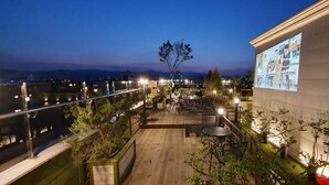 Eine Dachterrasse mit Außenmöbeln, einem großen Bildschirm, der Inhalte zeigt, und Blick auf die Stadt bei Dämmerung.