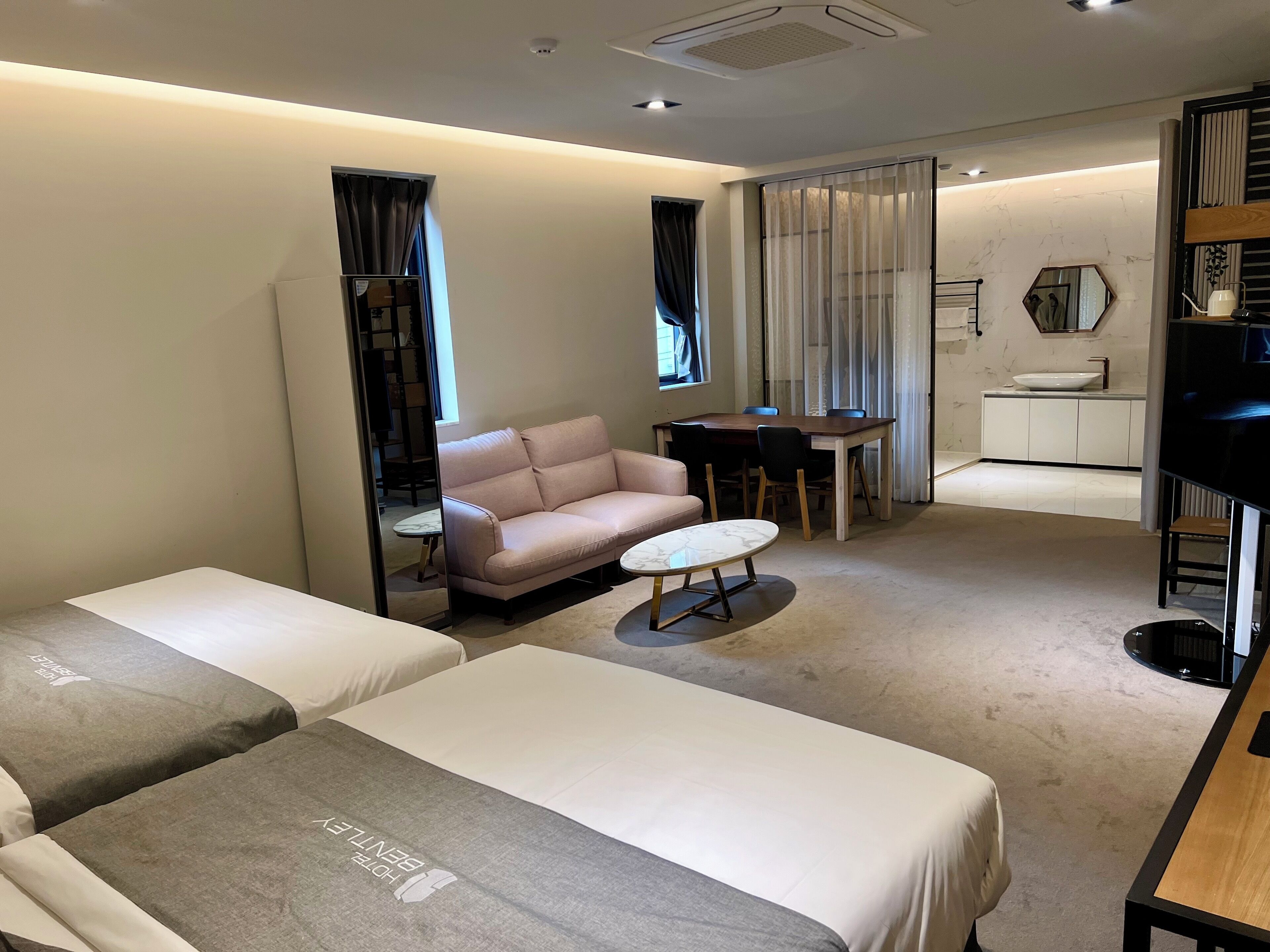 A hotel room with two beds, a sofa, a small table, a TV, and a bathroom with a sink and mirror.