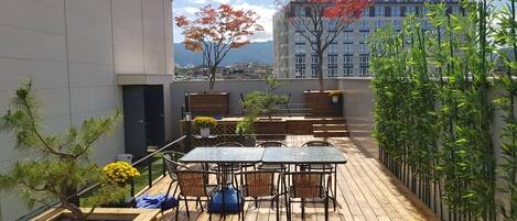 A rooftop patio with a wooden deck, a table with chairs, a small tree, and a bamboo fence.