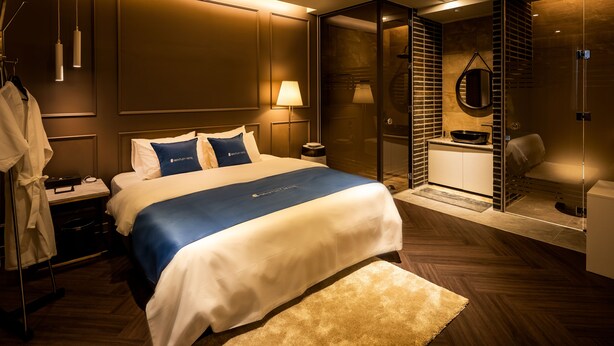 Ein modernes Hotelzimmer mit einem großen Bett, einem begehbaren Kleiderschrank, einem Badezimmer mit Glasdusche und Blick auf die Stadt.