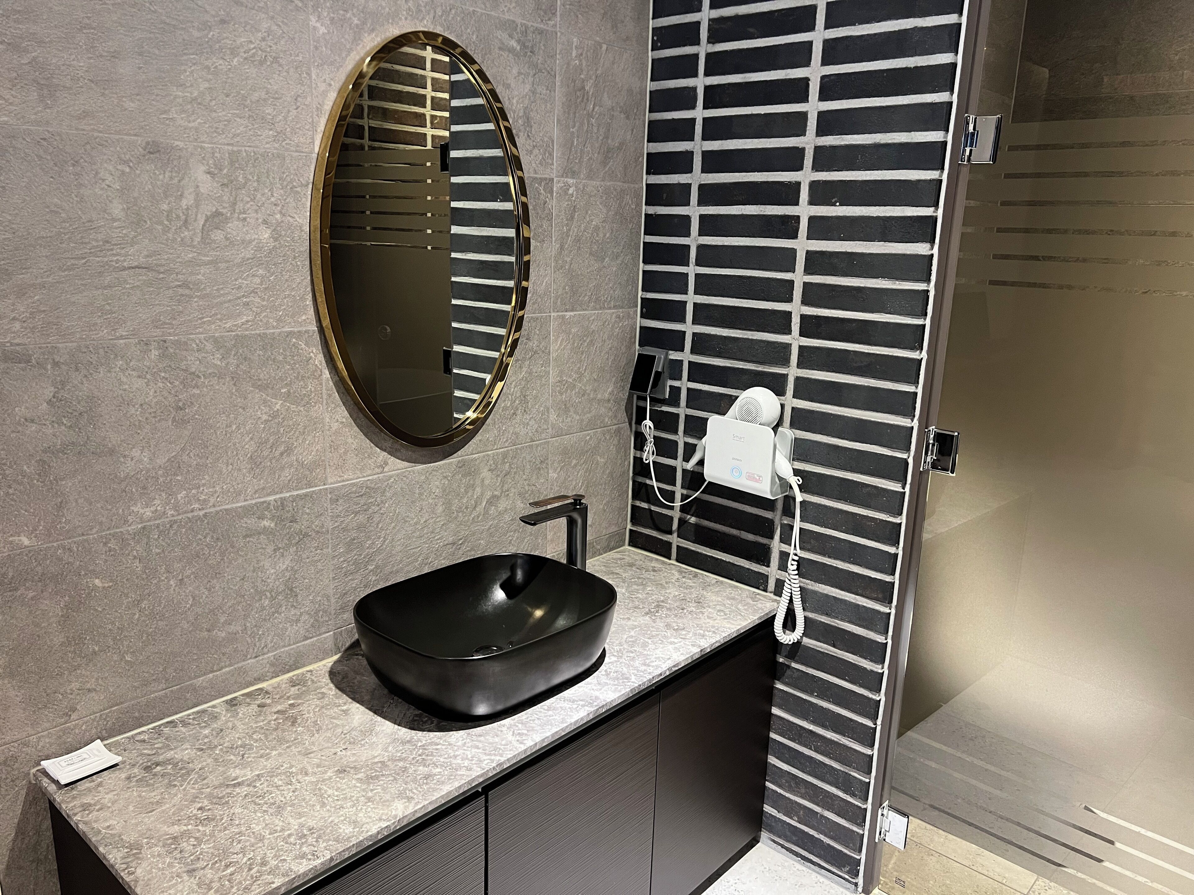 A bathroom with a black sink, a round mirror, and a hairdryer on a wall-mounted rack.
