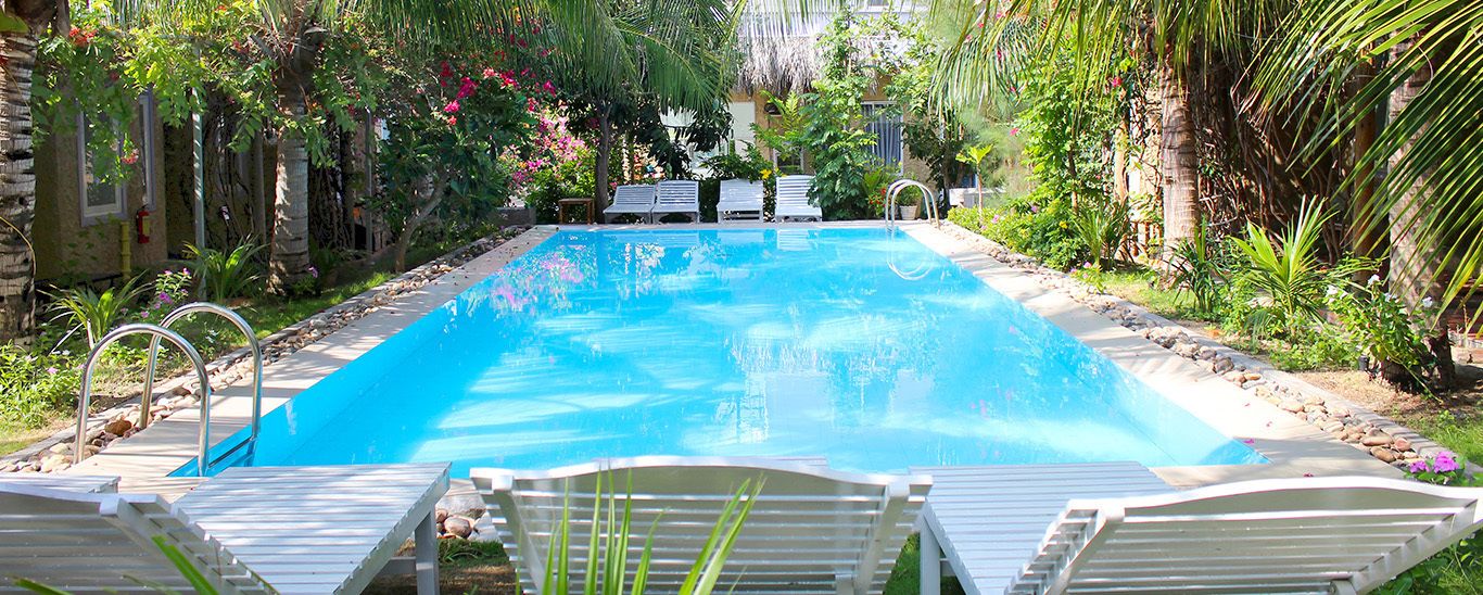 A rectangular swimming pool with a clear blue water surface, surrounded by lush greenery and palm trees. There are lounge chairs and a small table near the pool's edge.