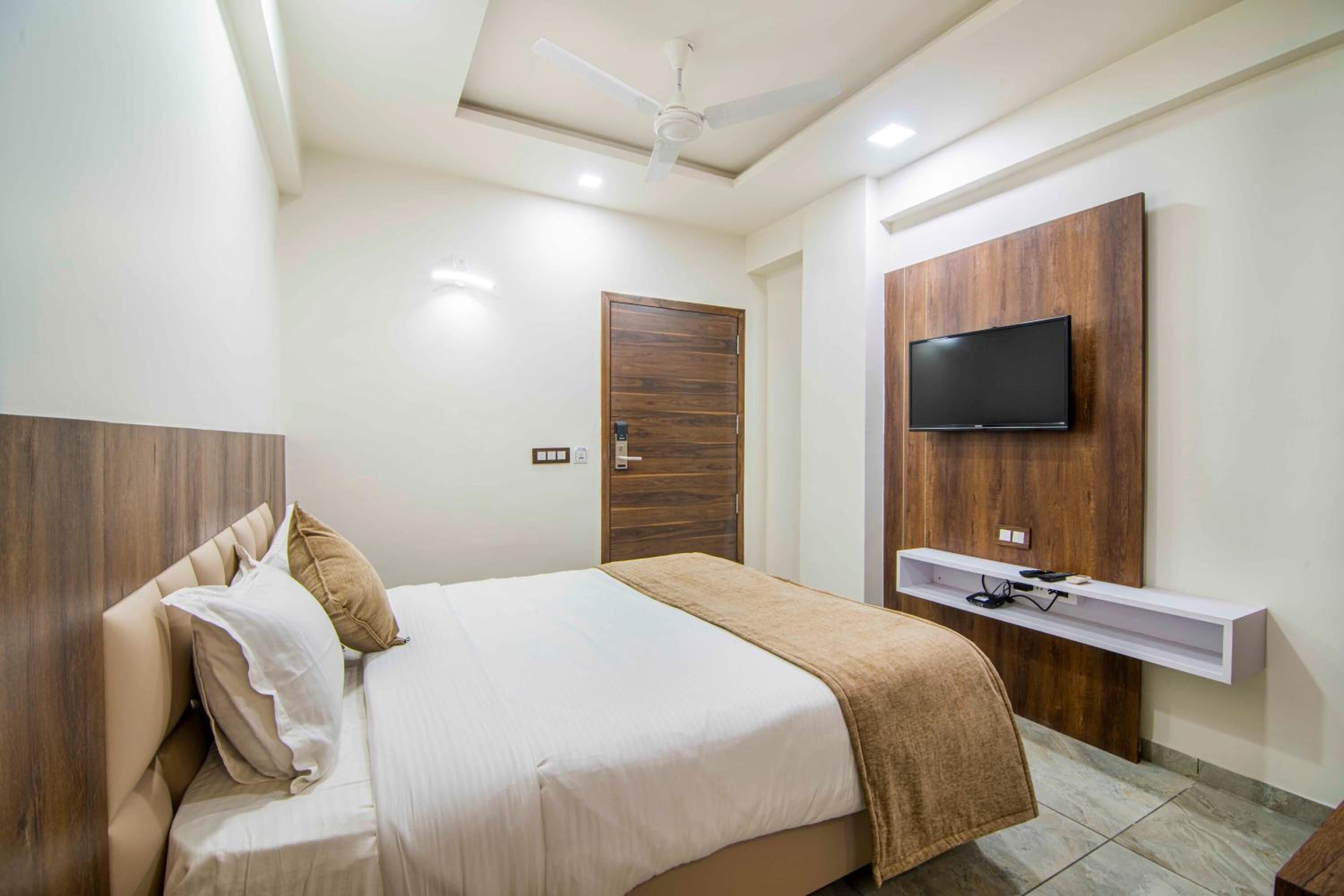 Un dormitorio con cama, ventilador de techo, televisor montado en un panel de madera y un estante pequeño debajo del televisor.