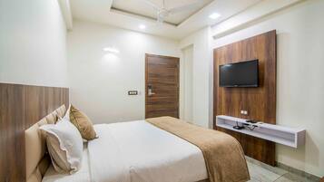 Un dormitorio con cama, ventilador de techo, televisor montado en un panel de madera y un estante pequeño debajo del televisor.