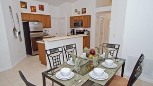 A dining area with a glass table set for four, surrounded by chairs. A kitchen with wooden cabinets and a microwave is visible in the background. - Sm218320 - Vizcay - 3 Bed 2 Baths Villa (Davenport)