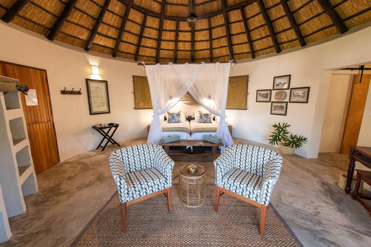 A room with a thatched roof, a bed with a canopy, two patterned chairs, a small table, and framed pictures on the walls.