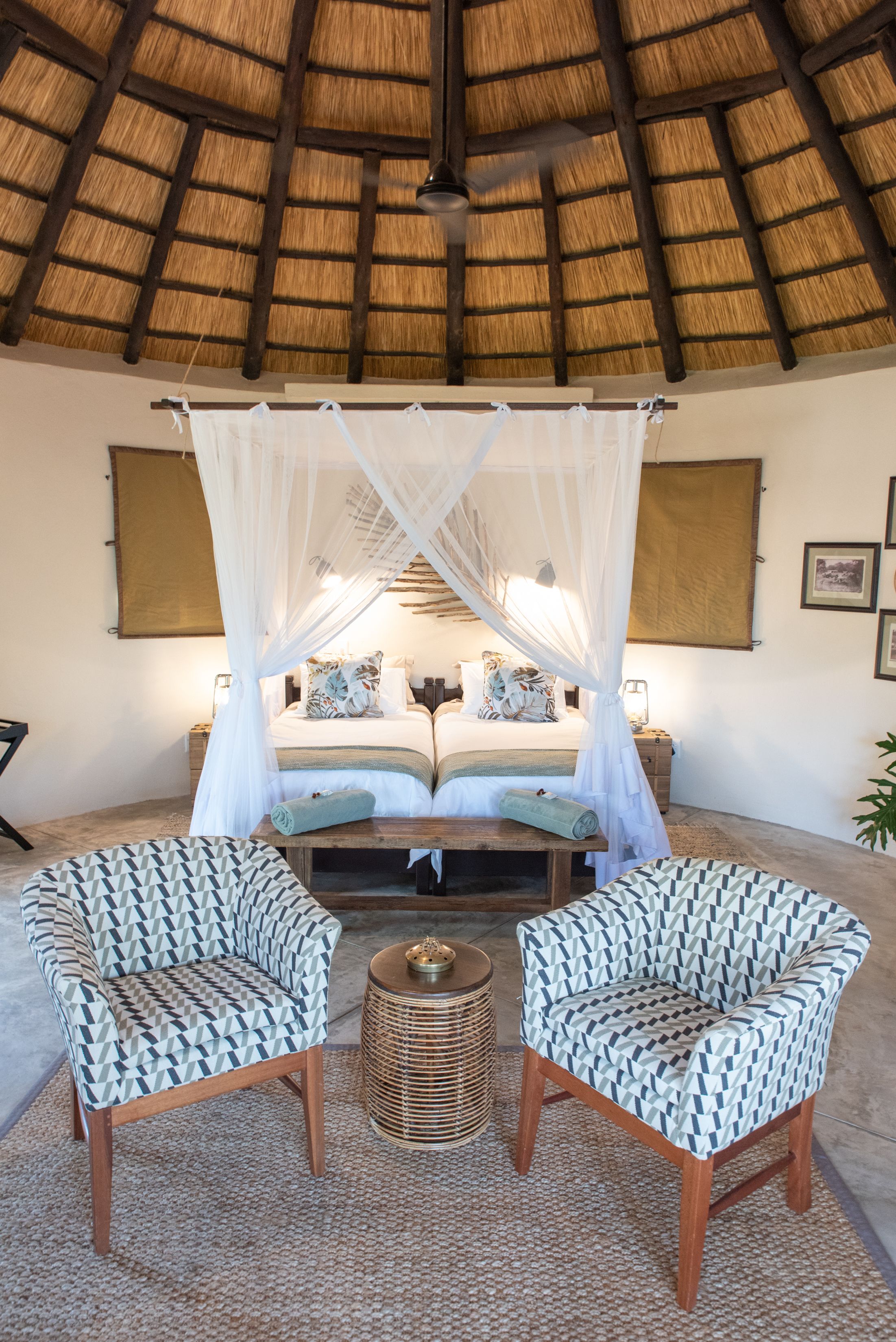 Une chambre à coucher avec un toit de chaume, un lit à baldaquin, deux fauteuils à motifs, une table ronde en bois et un panier tressé.