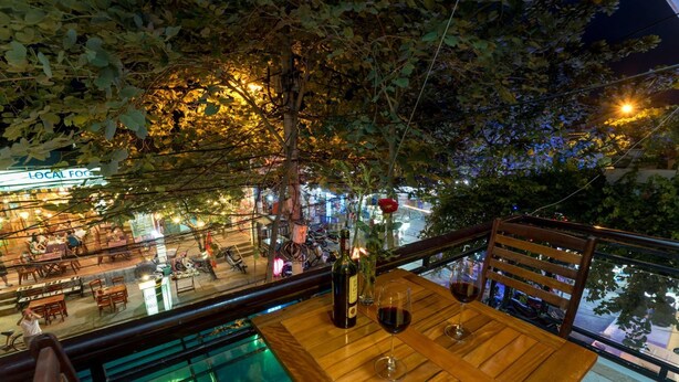 Una mesa de madera con dos copas de vino y una botella, frente a un animado ambiente de calle y un restaurante.