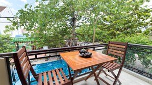 Un balcón con una mesa y sillas de madera, una piscina pequeña y vistas a árboles y edificios.