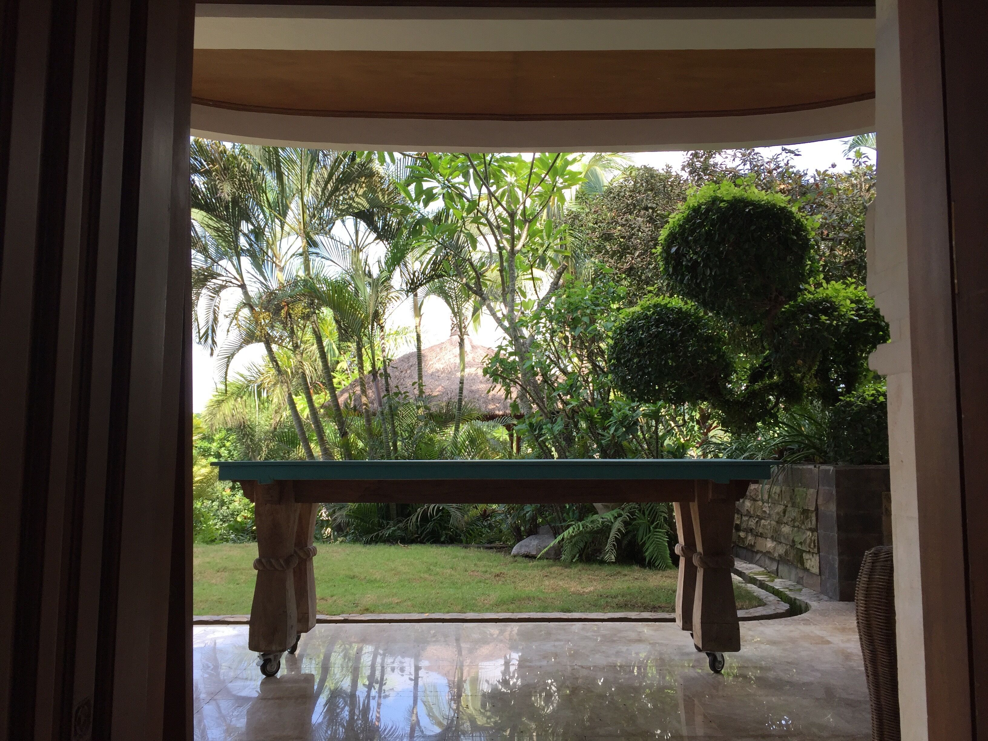 Visão através de uma porta para um pátio com mesa e cadeiras, cercado por vegetação exuberante e uma parede de pedra.