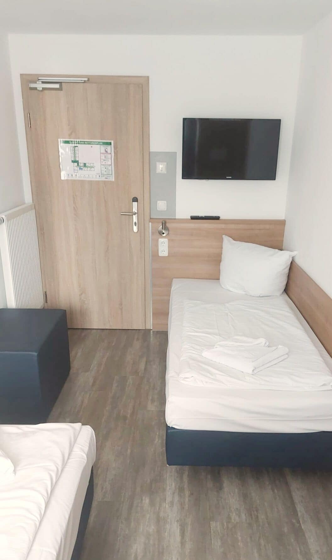 Hotellihuone, jossa on sänky, seinälle asennettu televisio, ovessa oleva kartta ja pieni pöytä.