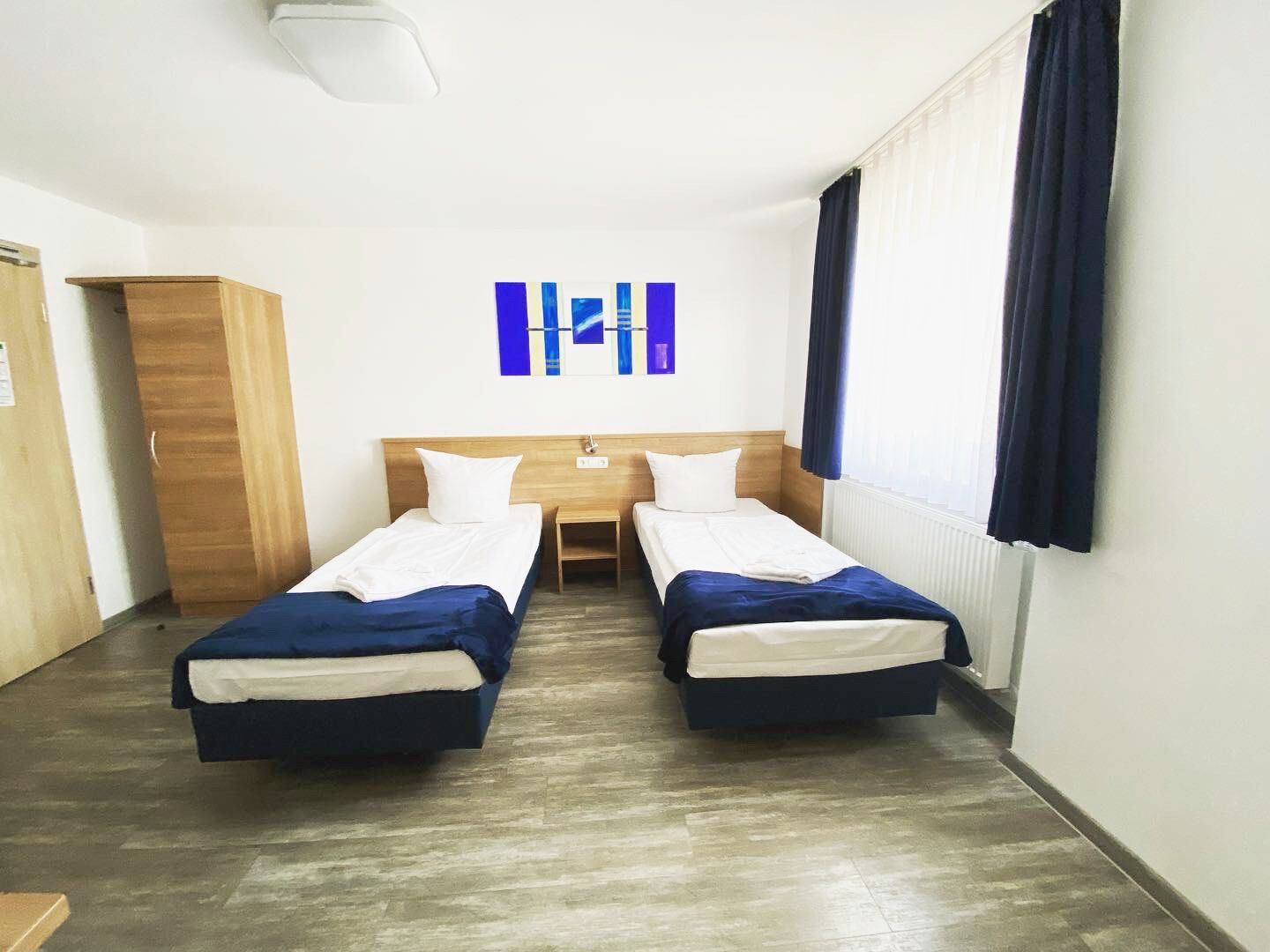Une chambre d’hôtel avec deux lits simples, une armoire en bois, une petite table et une œuvre d’art accrochée au mur.