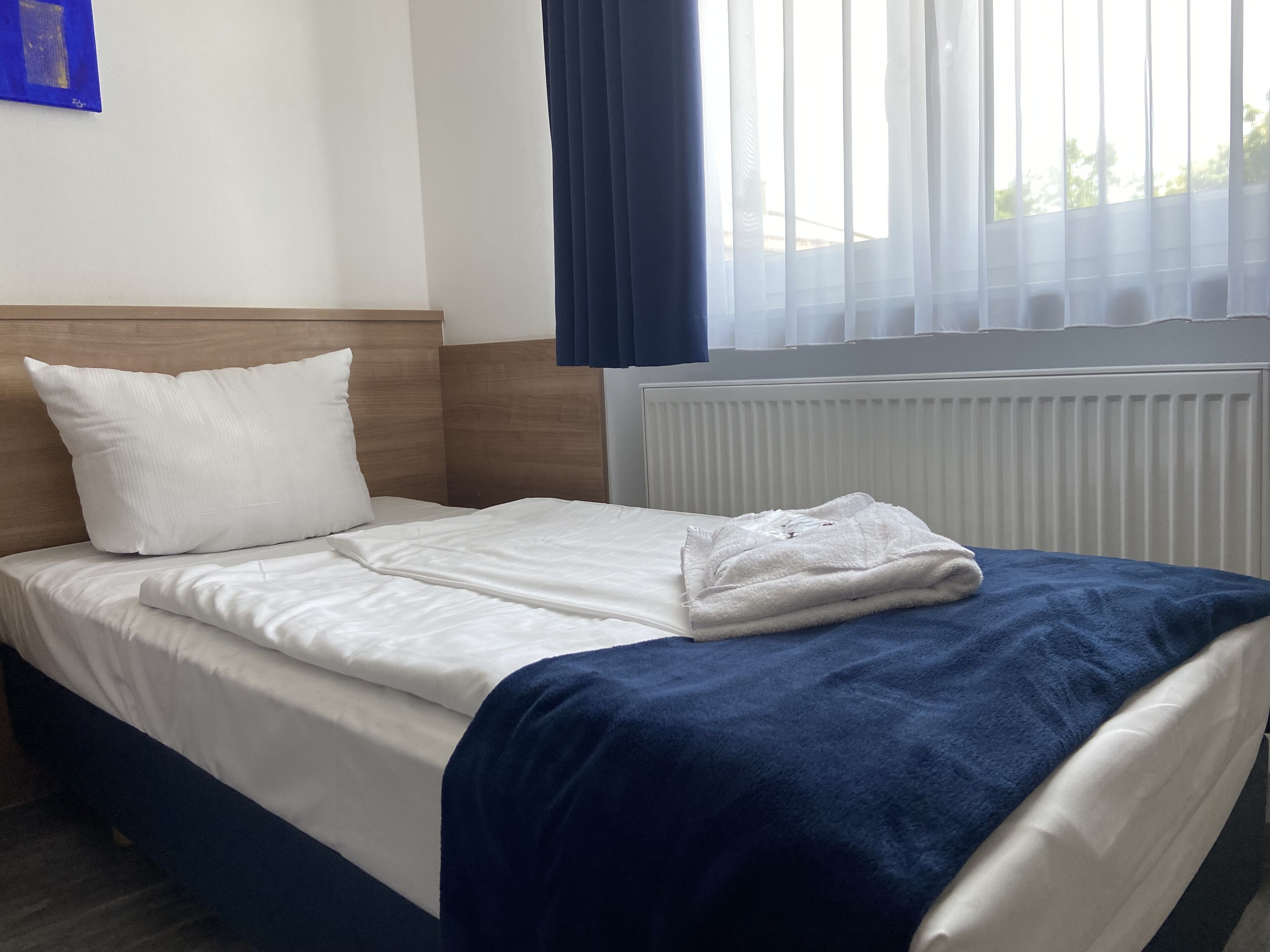 Een netjes opgemaakt bed met witte lakens en een blauwe deken, een houten hoofdbord en een witte radiator.