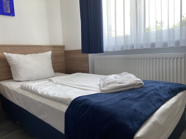 Un lit bien fait, avec du linge de lit blanc et une couverture bleue, une tête de lit en bois et un radiateur blanc.