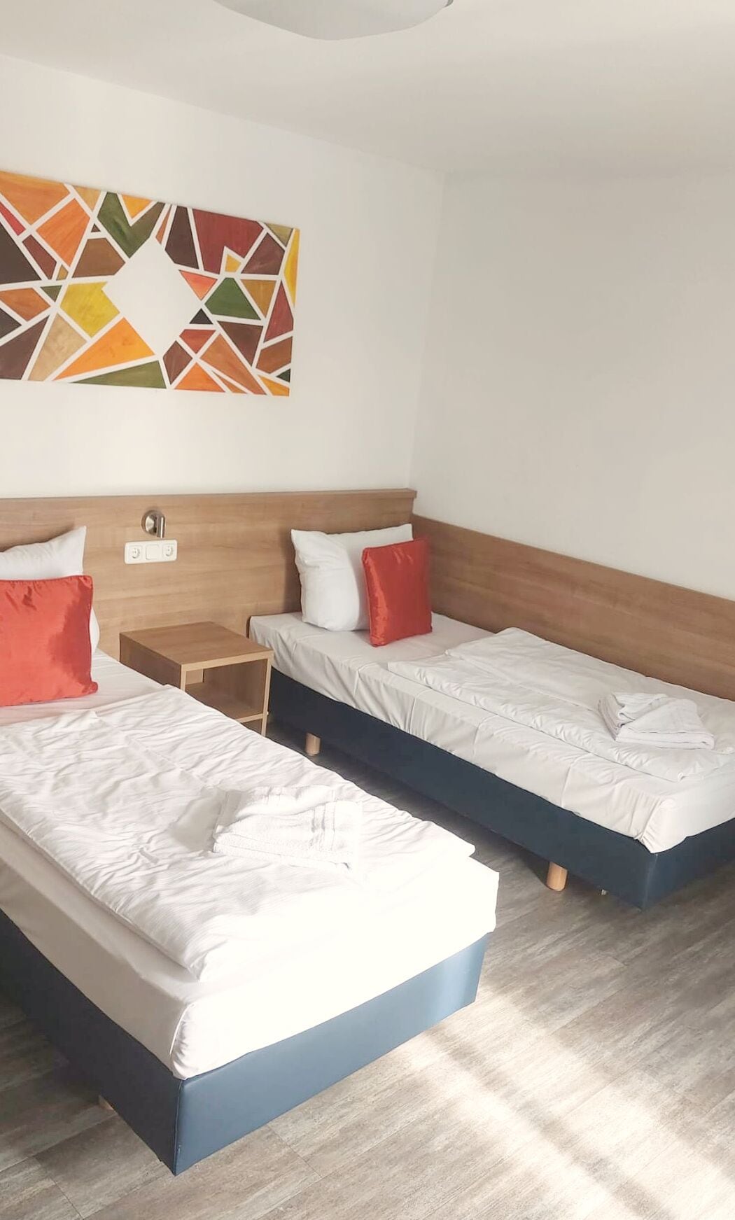 A hotel room with two beds, wooden headboards, white bedding, and a colorful geometric wall art piece.