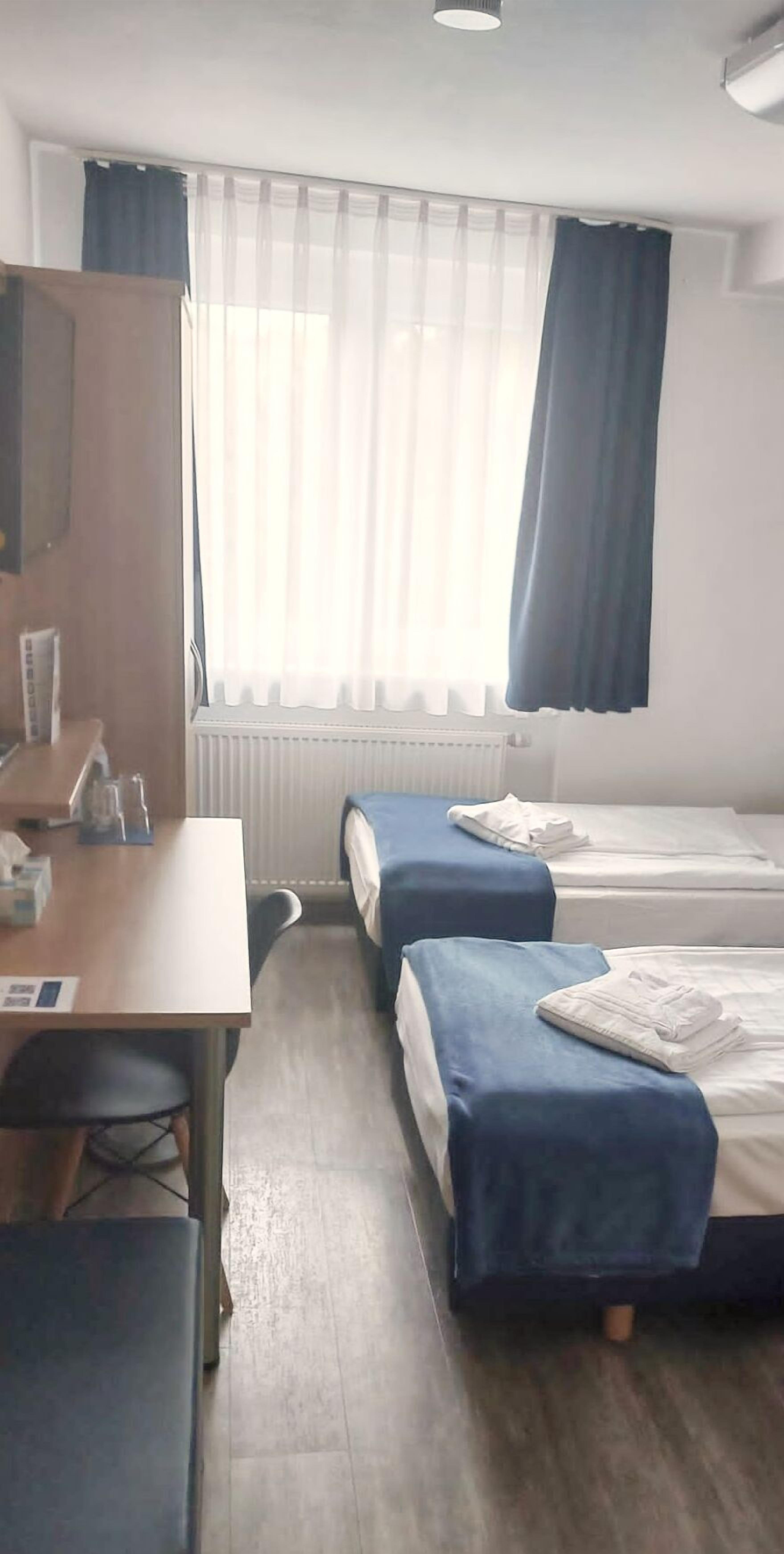 Habitación de hotel con dos camas individuales, un escritorio con silla, un radiador y una ventana con cortinas translúcidas.