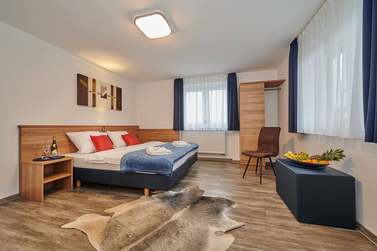 Une chambre d’hôtel moderne avec un lit en bois, un bureau, une chaise, une grande fenêtre avec des rideaux et un tapis en fourrure unique.