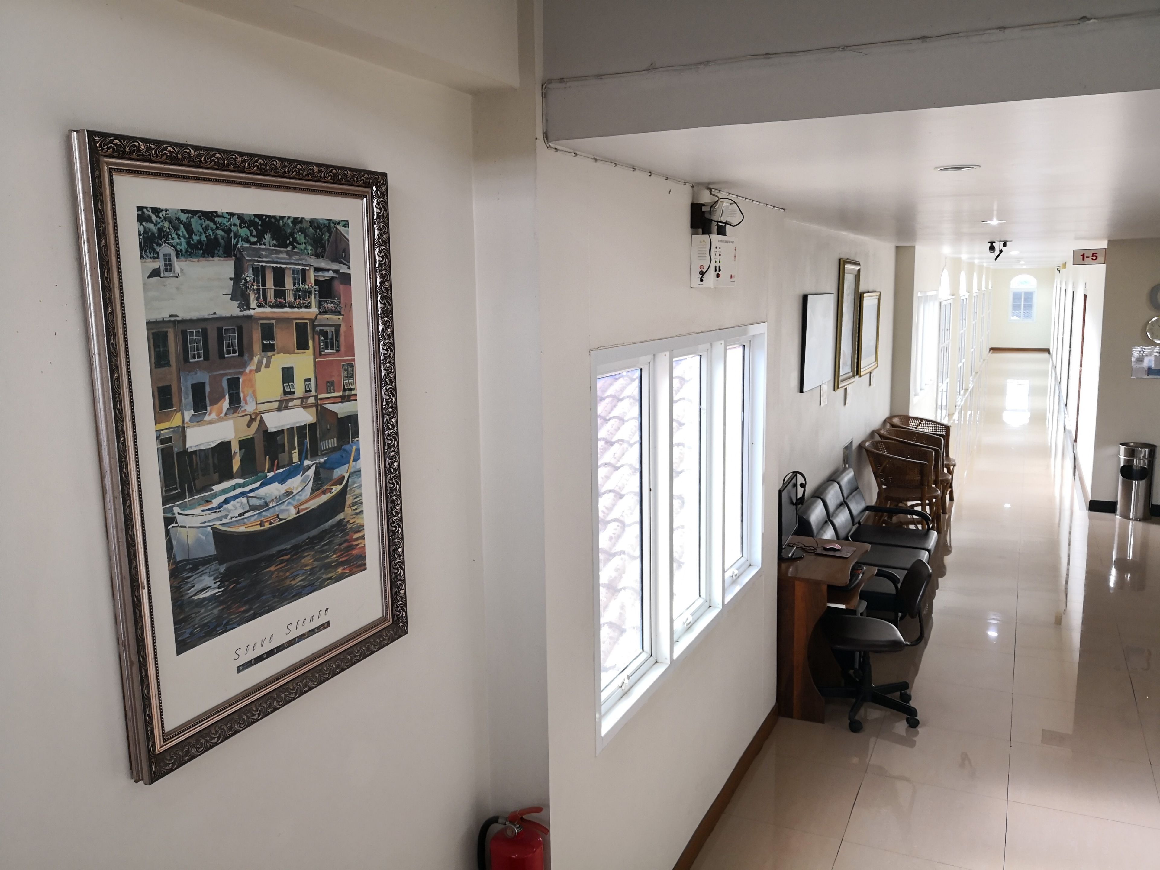 A hallway with a framed painting of a canal scene, a fire extinguisher, and a row of chairs along the wall.