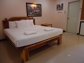 A neatly made bed with white linens and two towels, a wooden headboard, a small bedside table, and a framed painting on the wall.
