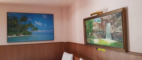 A hotel room with a bed, a painting of a beach, and a framed picture of a waterfall.