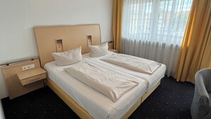 Quarto de hotel com cama de casal, mesa de cabeceira, luminária, cadeira e janela com cortinas.