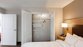 Habitación de hotel con cama bien tendida, mesita de noche con lámpara, armario con estantes y puerta que da a otra habitación.