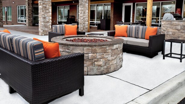 An outdoor seating area with wicker furniture and orange cushions, a stone fire pit, and a covered patio with a grill. - TownePlace Suites by Marriott Louisville Airport (Louisville)