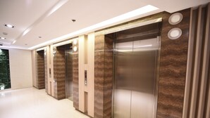 Área de elevador moderna com painéis metálicos e de madeira, chão limpo e teto com iluminação embutida.
