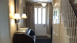 A hallway with a piano, two wall-mounted lamps, a chandelier, a door with curtains, and a staircase. - Carrick's At Castle Farm (Dereham)