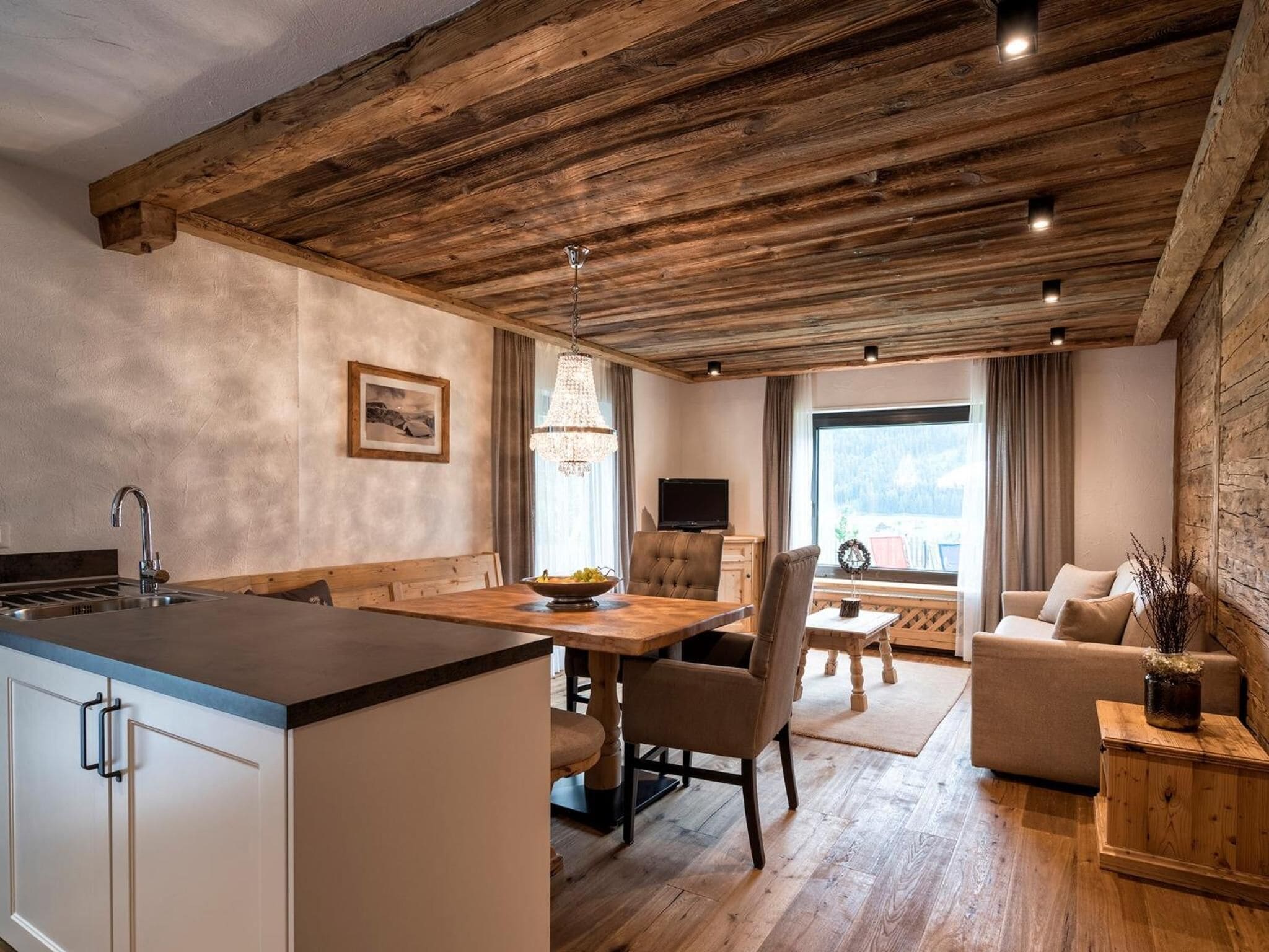 Une cuisine moderne avec un plafond en bois, un coin repas agrémenté d’un lustre, et un salon avec un canapé et un téléviseur.