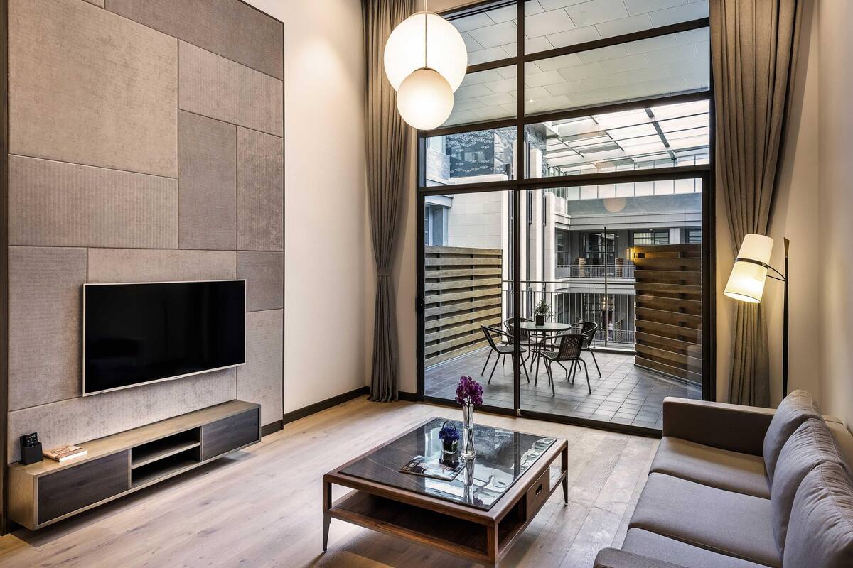 Una sala moderna con una televisión de pantalla plana, una mesa de centro de vidrio, un sofá y un área de comedor visible a través de amplios ventanales.