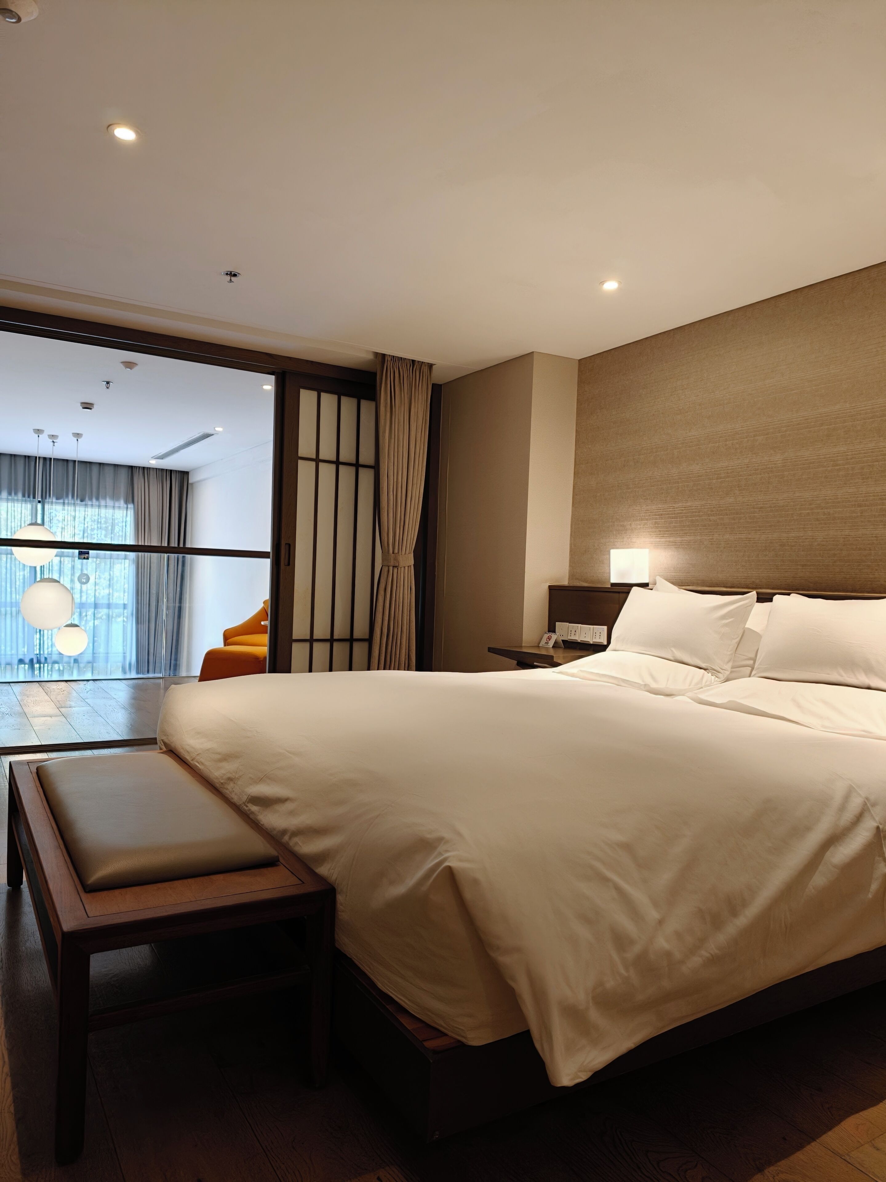 Habitación de hotel moderna con una cama grande, un banco, una mesita de noche con lámpara y vistas al exterior.