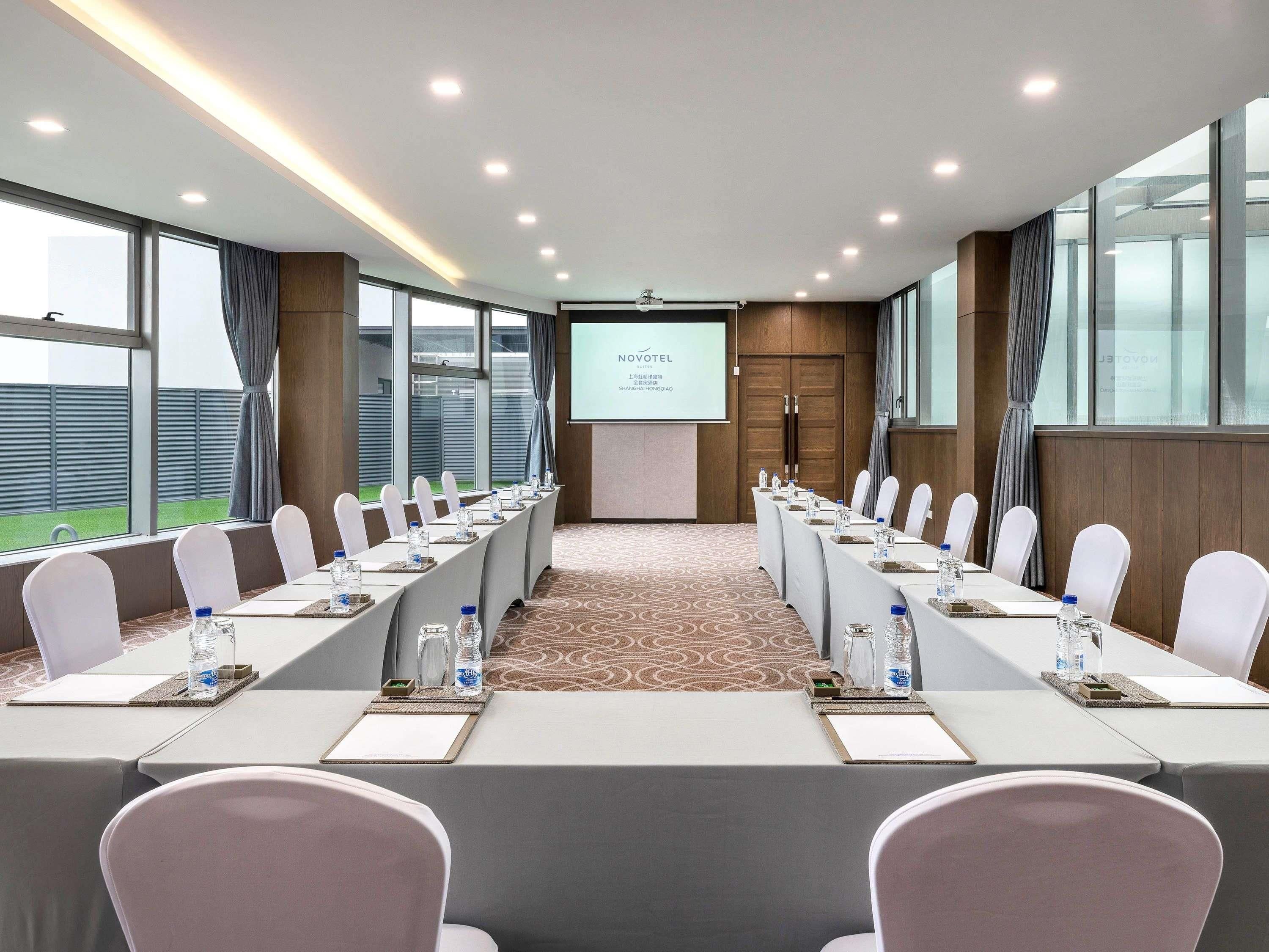 Un salón de conferencias con una larga mesa, sillas blancas, una pantalla de proyector y vistas a un edificio.
