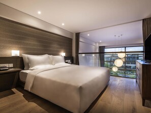 A modern hotel room with a large bed, bedside tables, a desk, and a view of the city through large windows.