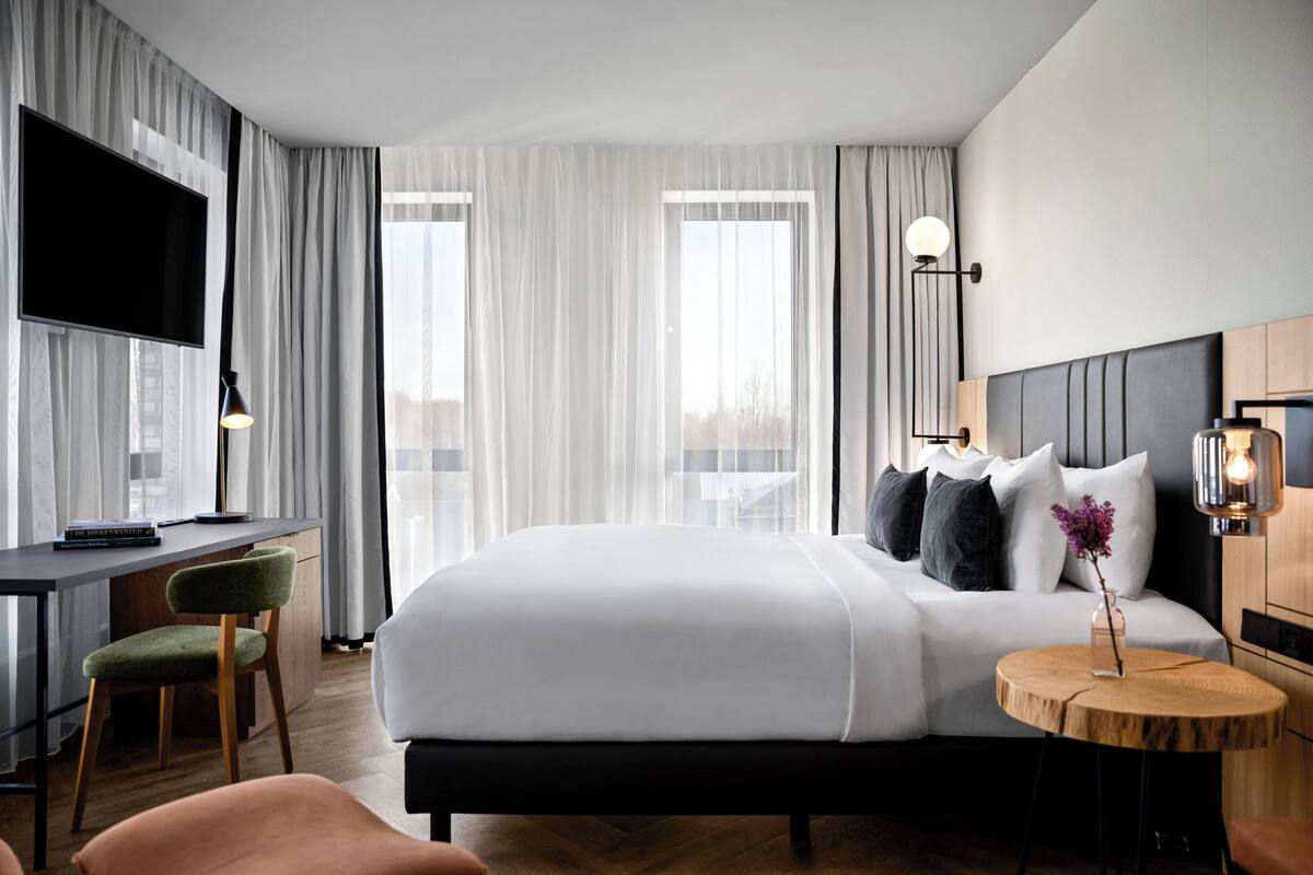 Habitación de hotel moderna con una cama grande, un escritorio con silla, un televisor y una mesa redonda de madera con un florero.