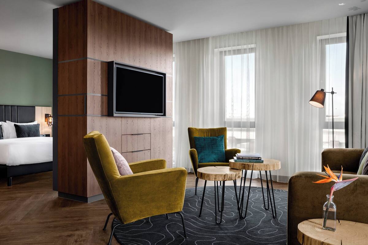 Una habitación de hotel moderna con una cama, dos sillones, una pequeña mesa redonda, un televisor montado en un panel de madera y amplias ventanas con cortinas translúcidas.