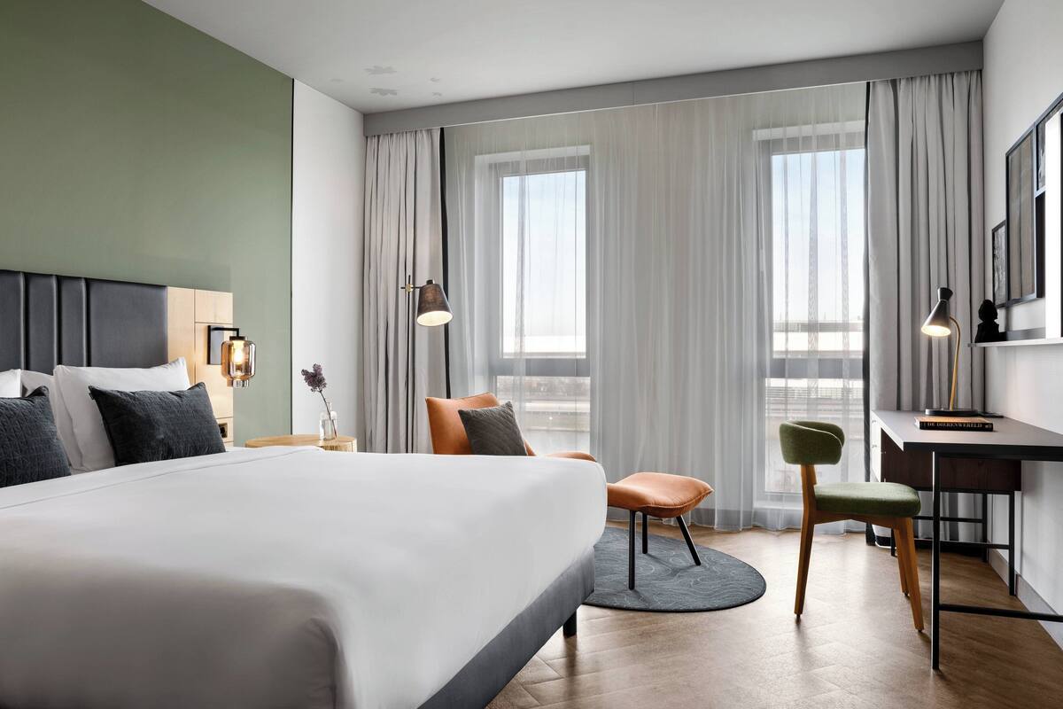 Una habitación de hotel moderna con una cama grande, un escritorio y una silla. Hay dos ventanas con cortinas translúcidas y vistas a la ciudad.